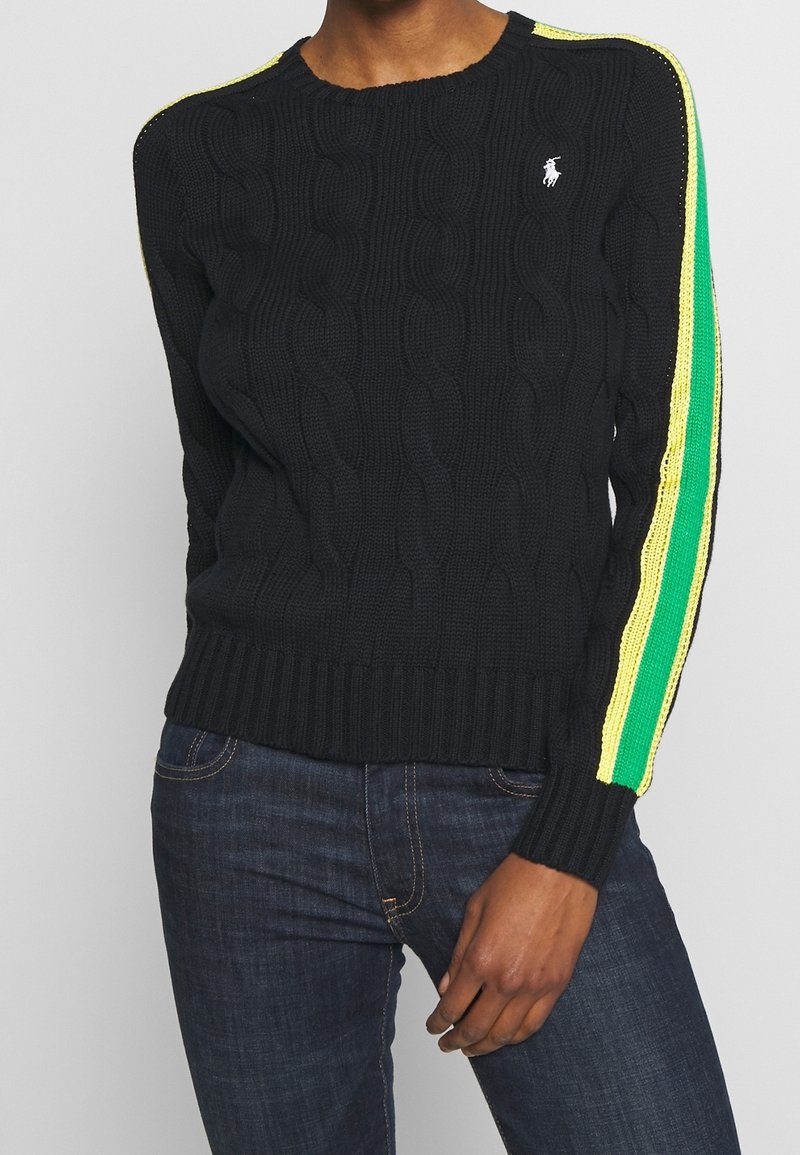 Svart kabelstickad tröja med ribbad nederkant, gula och gröna randiga detaljer på ärmarna och en liten logotyp på bröstet.