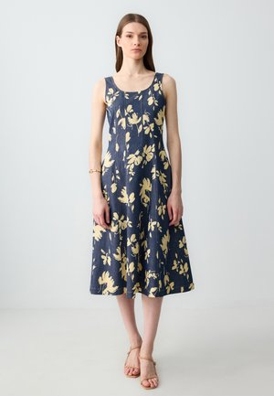 Vrouw die staat in een mouwloze marineblauwe midi-jurk met beige bloemmotief en beige sandalen met hakken tegen een effen lichte muur.