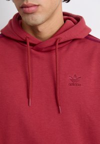 Rdeča bombažna kapuca z vrvico za zategovanje, ki ima subtilno vezeno logo na sredini. Gladka tekstura z minimalisticnim dizajnom.