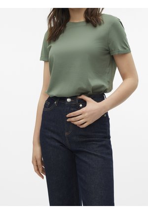 Kvinde iført en grøn kortærmet t-shirt stoppet ned i højtaljede mørkeblå jeans med den ene hånd hvilende nær linningen.