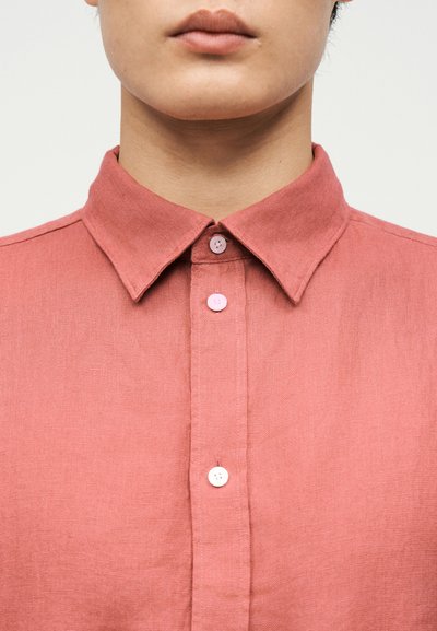 Κοντινό πλάνο του κάτω μέρους του προσώπου και του λαιμού ενός ατόμου που φοράει πουκάμισο από λινό σε σκόπινο ροζ χρώμα με κουμπιά και μυτερό γιακά.