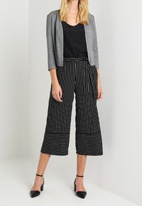 Blazer grigio sopra una maglietta nera, abbinato a pantaloni cropped a righe bianche e nere. Scarpe nere a punta, con una texture liscia.