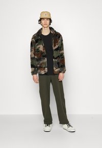 Fleecejakke med et multifarvet camouflage mønster, sort t-shirt, grøn fleecebukser og hvide sneakers. Beige cap med logo detaljer.