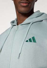 Φούτερ με φερμουάρ σε ανοιχτό πράσινο, κατασκευασμέν ο από μαλακό ύφασμα, με κεντημένο λογότυπο τριών ρίγων στο στήθος και κορδόνια στη κουκούλα.