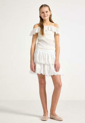 Ung pige står iført en hvid off-shoulder top og matching hvid øjet spidsnederdel med beige blomstrede balletsko, mod en ensfarvet baggrund.