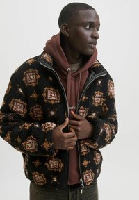 Giacca in pile nera con un motivo geometrico marrone e crema, dotata di chiusura con zip e una consistenza morbida, indossata sopra una felpa bordeaux.