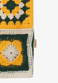 Tissu crocheté présentant des motifs géométriques verts, jaunes et blancs avec une petite étiquette de marque visible sur le côté droit.