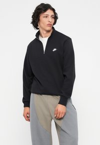Fekete, cipzáros pulóver, pamut keverékből készült, fehér Nike logóval a mellen. A modellen szürke, textúrázott, keskeny szárú nadrág van.