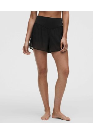 Femme portant un short en maille noire à taille haute et à plusieurs couches, debout pieds nus contre un fond clair uni.