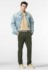 Giacca di jeans azzurro chiaro sopra un maglione beige, pantaloni verde oliva con una cintura nera e bianca a motivi, e sneakers beige.