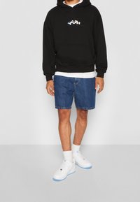 Felpa nera con cappuccio con logo "YOUTH" bianco e blu, abbinata a pantaloni corti in denim blu e sneakers bianche con accenti blu chiaro.