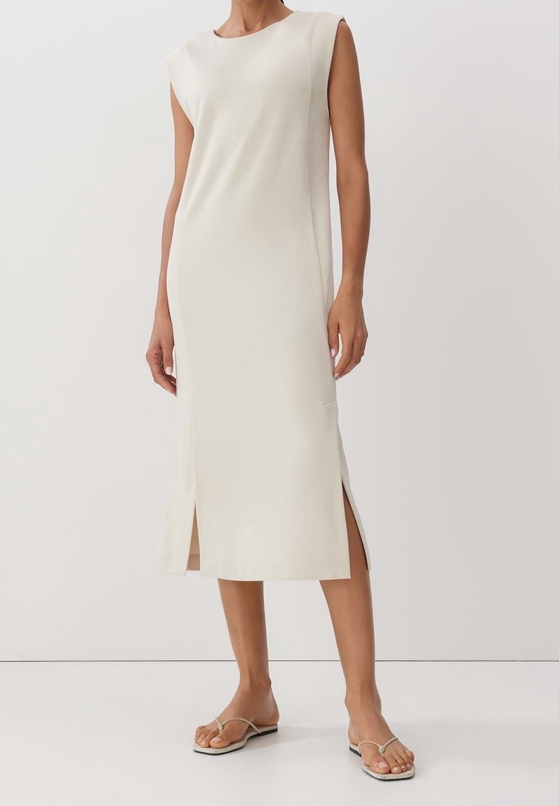 Kobieta w niebieskiej, beżowej sukience midi bez rękawów z bocznymi rozporkami i płaskich beżowych sandałach na płaskiej podeszwie, stojąca na jednolitym białym tle.