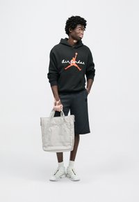 Μαύρο φούτερ με κουκούλα με λογότυπο "Air Jordan" σε λευκό και πορτοκαλί, γκρι σορτς, κρατώντας μια ανοιχτό γκρι, μαλακή τσάντα ώμου. Λευκά αθλητικά παπούτσια με λεπτομέρειες.