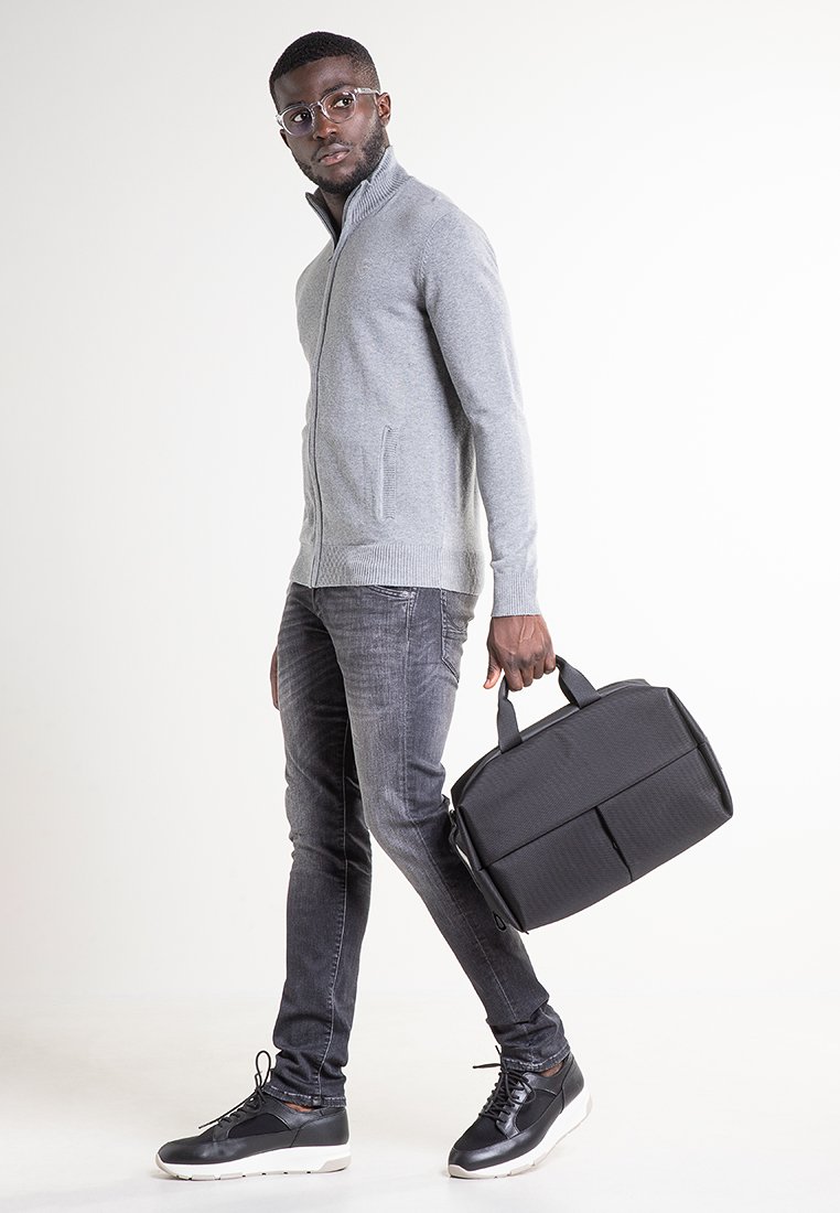 Pull gris à fermeture éclair, jean foncé et baskets noires. Tient un sac rectangulaire noir à deux compartiments, avec des poignées robustes.