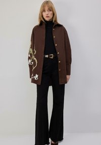 Pruun nööpidega särk kraega, mille vasakul varrukal on rohevalge lillemustriga tikand, koos mustade lehvikukujuliste pükstega.