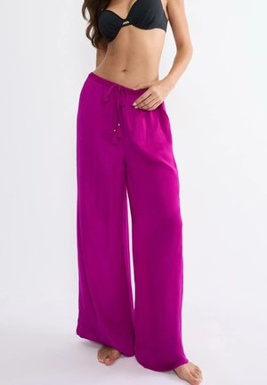 Kobieta w czarnym topie bikini i luźnych, jasnoróżowych spodniach z szerokimi nogawkami stojąca boso na białym tle.