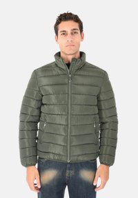 Giacca verde imbottita con colletto alto, trapuntatura orizzontale, tasche con zip e una texture liscia e lucida. Indossata sopra una maglietta nera.