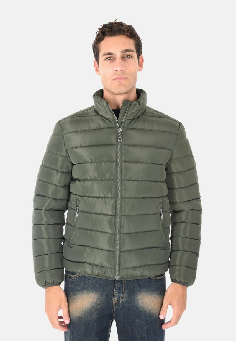 Giacca verde imbottita con colletto alto, trapuntatura orizzontale, tasche con zip e una texture liscia e lucida. Indossata sopra una maglietta nera.