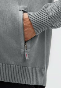 Grauer Strickpullover mit einer Reißverschlusstasche mit silbernen Beschlägen, gerippten Bündchen und einer strukturierten Oberfläche. Der Stoff wirkt weich und vielseitig einsetzbar.