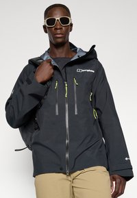 Zwarte waterdichte jas met grijze voering, groene ritsaccenten, meerdere voorkruimelzakken en logo. Gekocht met lichtbeige broek en zonnebril.