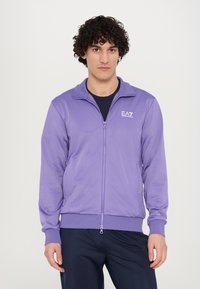 Violetas zip-up jaka ar augstu apkakli un garām piedurknēm. Materiāls izskatās gluds un nedaudz spīdīgs. Krūtīs ir logo.