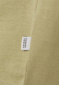 Světlý zelený bavlněný materiál s jemnou texturou. Obsahuje bílou štítek přišitou s nápisem "FRIDAY CASUAL." Jednoduchý a moderní design.