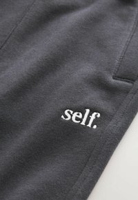 Šedé džínové kalhoty s malým bílým vyšitým logem "self.", hladkou texturou a minimalistickým designem.