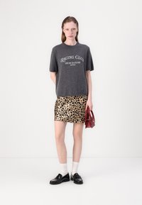Grauer Kurzarmpullover mit dem Text "RAGING CLUB", kombiniert mit einem Minirock im goldenen Leopardenmuster. Schwarze Loafers und eine rote Handtasche runden das Outfit ab.