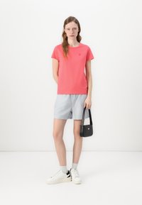 Koral kortermet t-skjorte, lysegrå shorts, hvite joggesko med svarte detaljer, og en liten svart håndveske. Enkelt stoff og avslappet passform.