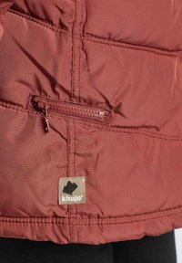 Gros plan sur une veste couleur rouille avec une poche zippée horizontale et un écusson beige de la marque Khujo près de l’ourlet.