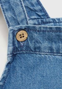 Denim kombinezoni z nastavljivimi naramnicami z lesenim gumbom in modrim šivom, prikazujejo srednje pranje tkanine.