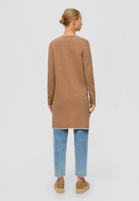 Pull en maille à manches longues de couleur brun clair, avec un col rond et des fentes sur les côtés, associé à un jean bleu clair et des chaussures à plateforme beiges.