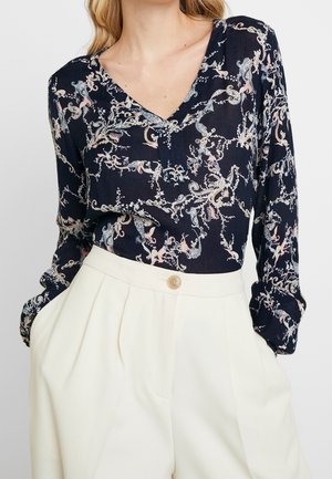 Woman wearing a navy V-neck blouse with a pastel floral pattern tucked into high-waisted white pleated trousers with a button closure.