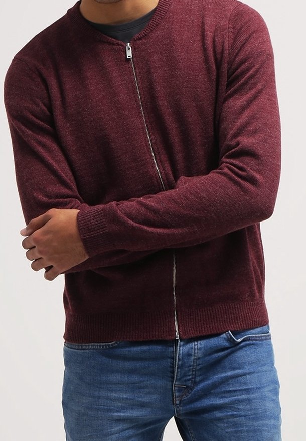 Homme portant un pull zippé bordeaux par-dessus une chemise noire et un jean bleu, les bras croisés devant un fond uni.