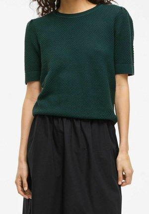 Kobieta ubrana w ciemnozielony, teksturowany, krótki sweter z dzianiny i czarną spódnicę z wysokim stanem, stojąca na jednolitym tle.