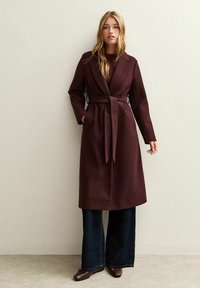 Cappotto avvolgente bordeaux con colletto a risvolto, vita con cintura e tasche a toppa. Abbinato a pantaloni in denim scuro a penna e scarpe coordinate.