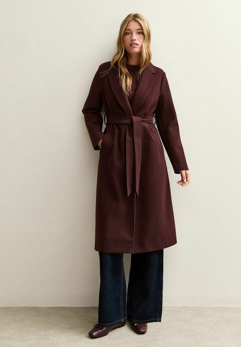 Cappotto avvolgente bordeaux con colletto a risvolto, vita con cintura e tasche a toppa. Abbinato a pantaloni in denim scuro a penna e scarpe coordinate.