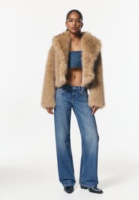 Veste courte en fausse fourrure marron clair, associée à un haut court bleu fonc é et un jean à jambes larges. Des bottines noires complètent la tenue.