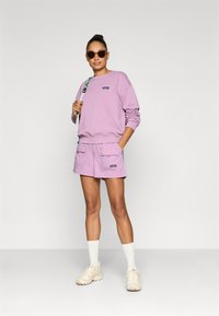 Μωβ φούτερ με ριμπ μανσέτες, σε συνδυασμό με μωβ σορτς που διαθέτουν δύο μπροστινές τσέπες. Το μοντέλο φορεί μπεζ αθλητικά παπούτσια και γυαλιά ηλίου.