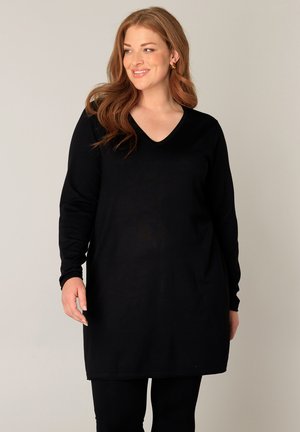 Lachende vrouw met lang rood haar, gekleed in een zwarte lange mouwen tuniek en een zwarte broek, staand tegen een effen achtergrond.