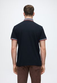 Laivastonsininen polo-paita lyhyillä hihoilla, jossa on punaiset ja harmaat raidat kauluksessa ja hihansuissa. Liukas rakenne, nappivapaa takasuunnittelu.