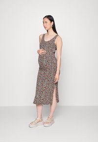 Vestido de maternidade em preto com um padrão floral que apresenta flores vermelhas e brancas. Design sem mangas, até ao joelho, com uma fenda lateral. Usado com sandálias de plataforma.