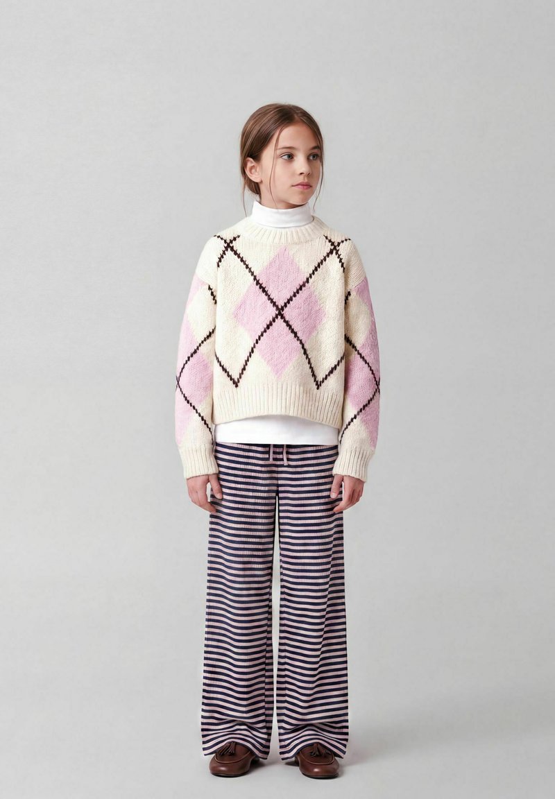 Jeune fille debout portant un pull argyle blanc et rose, un col roulé blanc, un pantalon large rayé et des chaussures marron sur un fond gris uni.
