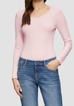 Nainen, joka pukeutunut vaaleanpunaiseen pitkähihainen V-paita, joka on sullottu sinisiin farkkuihin, toinen käsi taskussa yksiväristä taustaa vasten.