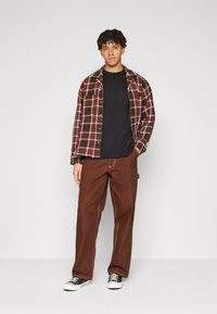 Brunternet flannel skjorte med knaplukning, sort t-shirt under, brune vide bukser og sorte sneakers med hvide såler.