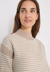 Beżowy sweter dzianinowy z prążkowanym kołnierzem, z wzorem w kratkę w jaśniejszych odcieniach, o miękkiej teksturze i opadających ramionach.