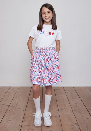 Ragazza giovane in piedi con le mani in tasca, indossa una maglietta bianca "ESCADA", gonna plissettata colorata, calzini bianchi e scarpe da ginnastica bianche su pavimento in legno.