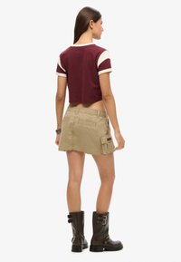 Korte maroon shirt met crèmekleurige accentstrepen, gecombineerd met een tan cargoschort met zijzakken en kniehoge bruine laarzen.