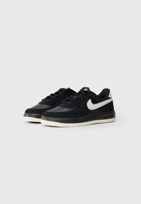 Par tenisice bez visine u crnoj boji s bijelim Nike swoosh logotipovima i bijelim potplatom na običnoj sivoj pozadini.