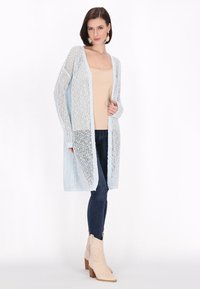 Cardigan azzurro aperto a maglia larga con maniche lunghe, texture leggera, abbinato a un top beige e jeans skinny scuri, e stivali da cowboy crema.